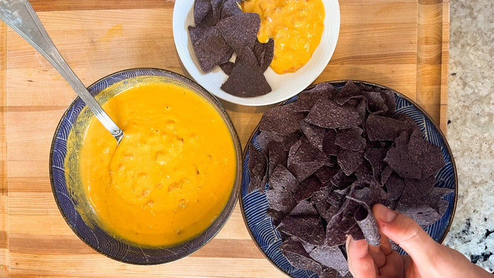 a hand picking up a blue corn tortilla chip next to a bowl of vegan cheese dip